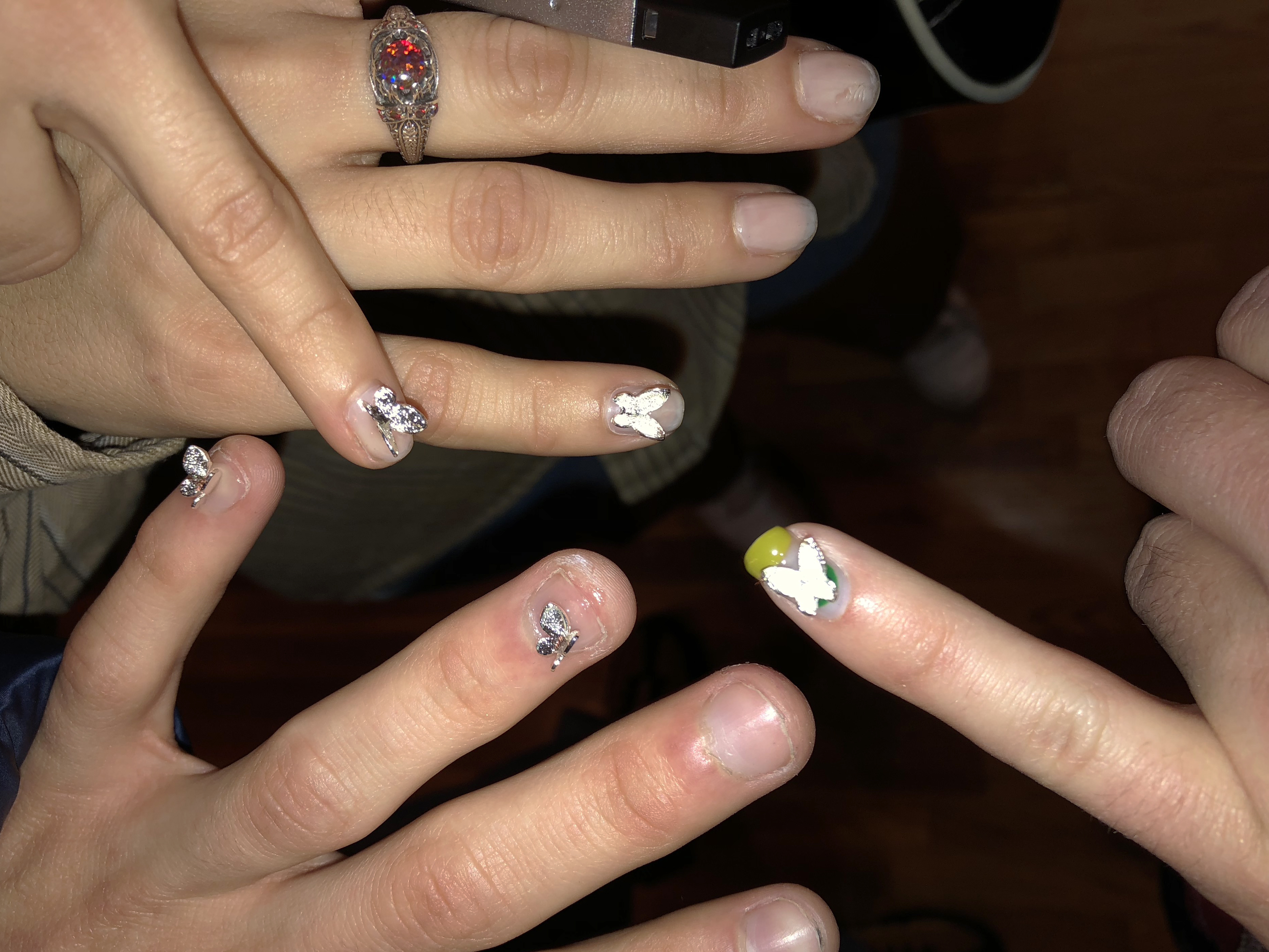 Four hands with fingers extended showing nails with butterfly charms adhered to them